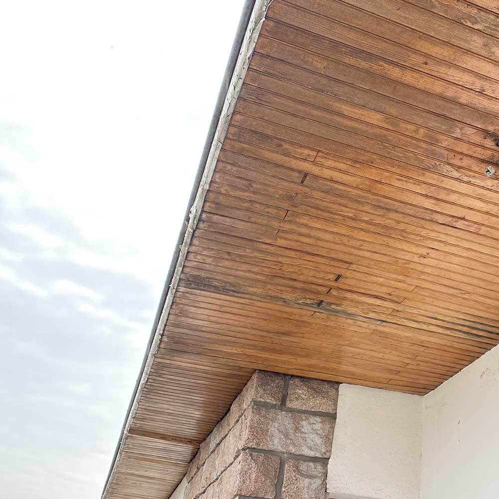 Sous-face de toit en bois : Entretien et rénovation Sous-face de toit en lambris de bois avec traces d'usure au-dessus d'un mur en pierre et façade blanche.